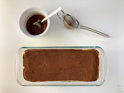 Tiramisu in a glass plate, cocoa powder in a white bowl, and tea strainer