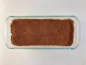 Tiramisu in a glass plate, cocoa powder in a white bowl, and tea strainer