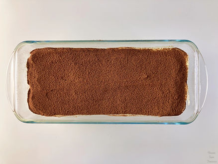 Tiramisu in a glass plate, cocoa powder in a white bowl, and tea strainer