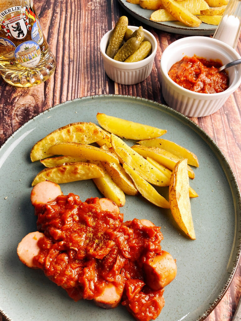 Grilled sausage, tomato curry sauce, french fries, salt, pickles and beer on a plate