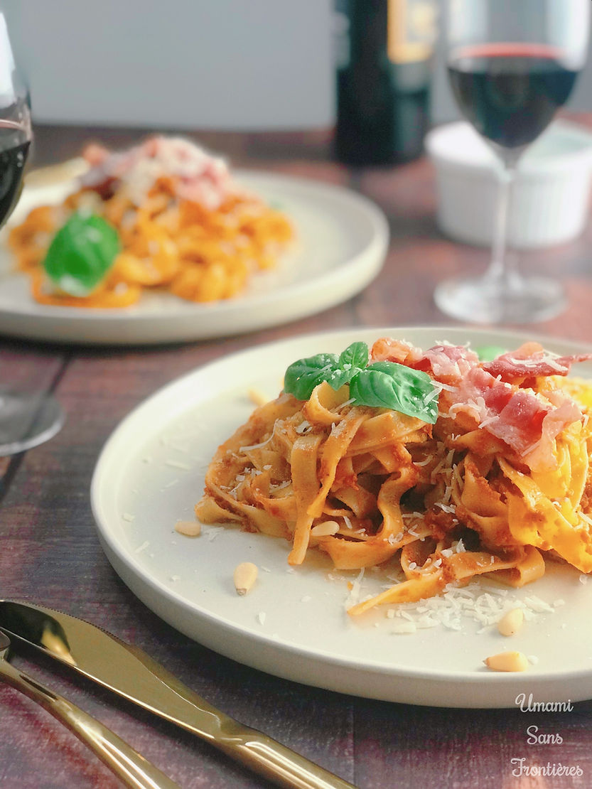 Bacon and pestosso pasta on a plate, pine nuts, wine glass, red wine, knife, fork, wine bottle and basil leaves