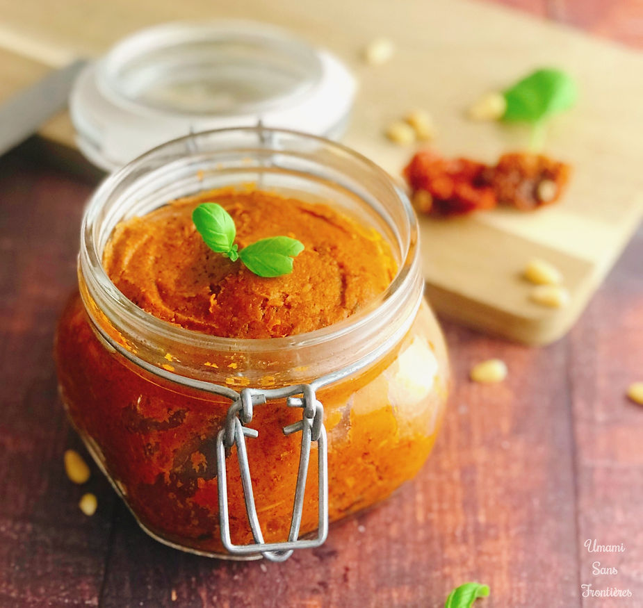 Sun-dried tomato pestosso in a glass container, knife, wooden board, sun-dried tomatoes, pine nuts and basil leaves