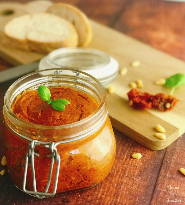 Sun-dried tomato pestosso in a glass container, knife, wooden board, sun-dried tomatoes, pine nuts, basil leaves, bread with sun-dried tomato pestosso, parmesan cheese